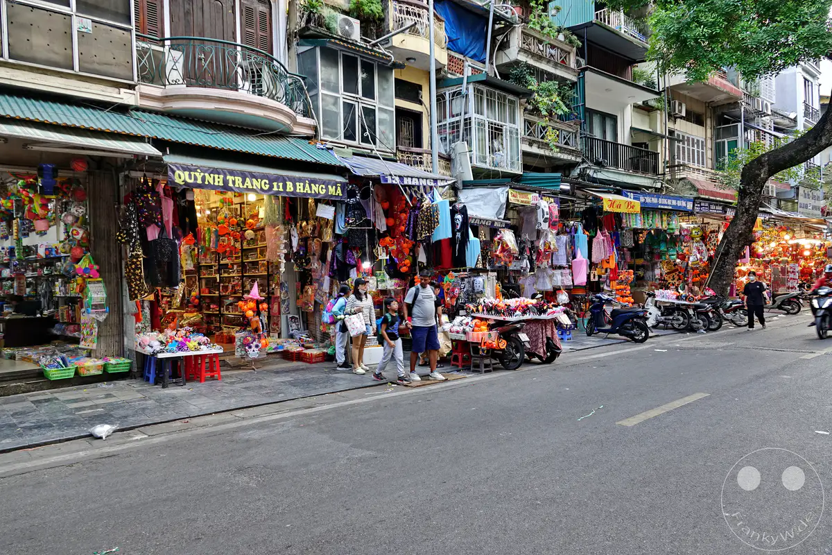 In Hanoi's Old Quarter – die Altstadt – säumen sich kleine Geschäfte, die mit bunten Souvenirs, Kostümen und Spielzeug gefüllt sind. Die belebte Einkaufsstraße lädt zum Stöbern ein, während Motorroller auf dem Asphalt vorbeiziehen. Eine lebhafte Atmosphäre herrscht zwischen den dicht gedrängten Ständen und den mehrstöckigen Gebäuden.