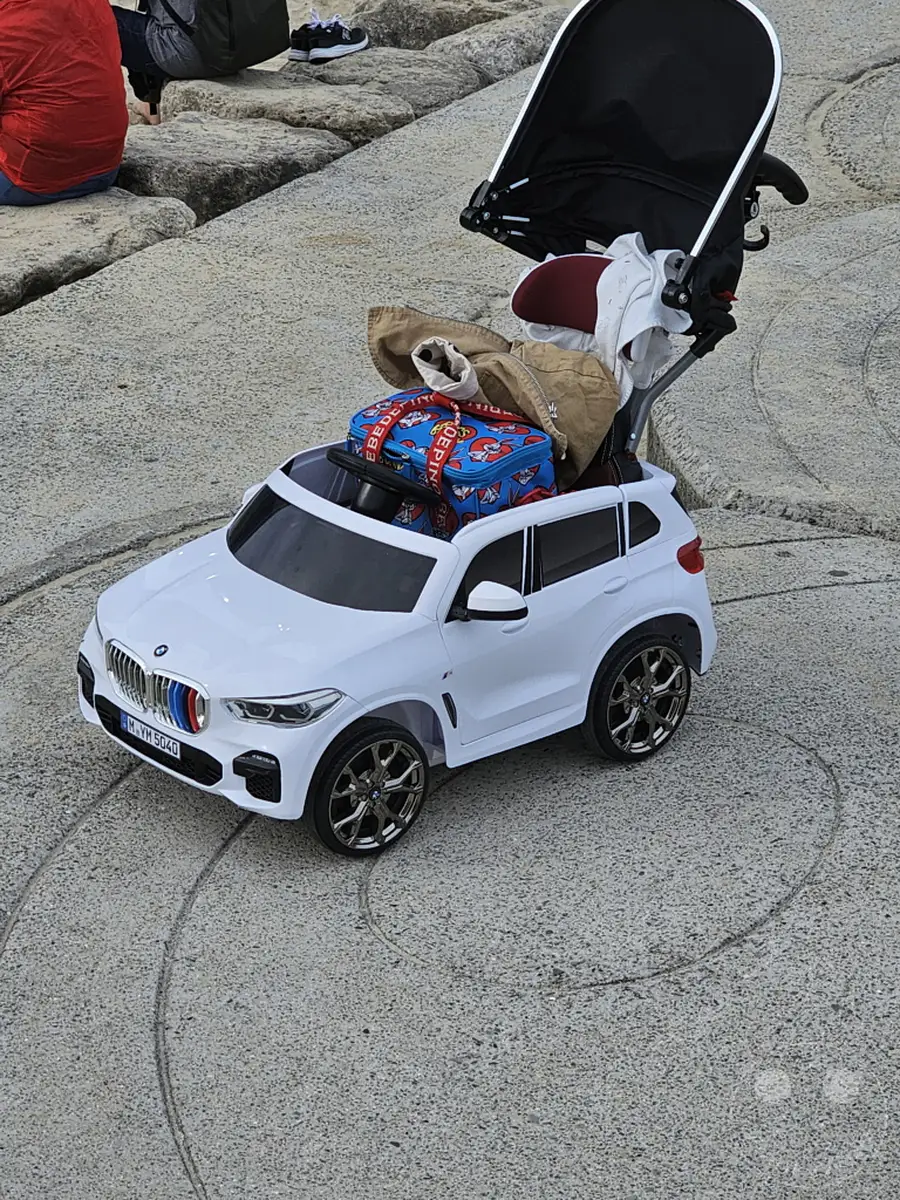 Ein weißes Kinder-BMW-Spielzeugauto ist mit einem Kinderwagen und Gepäck beladen, bereit für eine Ausfahrt am Haeundae Beach in Busan, Südkorea. Die Szenerie strahlt eine entspannte Atmosphäre aus, die auf einen Familienausflug oder Urlaub hindeutet. Die Steinoberfläche und die weite Fläche lassen auf einen öffentlichen Platz oder eine Promenade schließen.
