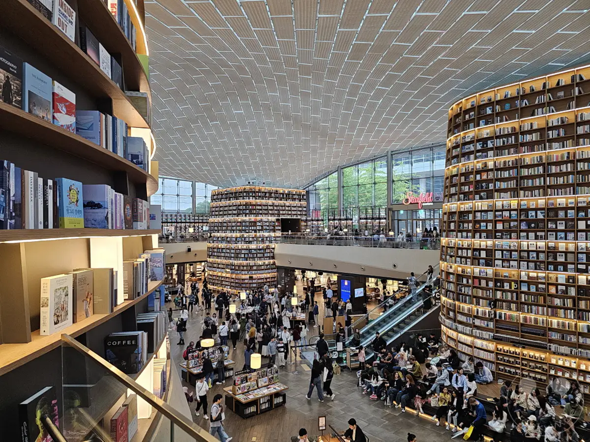 In Südkorea, Seoul, lädt die Starfield Library zu einem Besuch ein, wo hohe Bücherregale eine beeindruckende Architektur präsentieren. Menschenmassen verteilen sich zwischen den Gängen, viele sind tief in Bücher vertieft oder unterhalten sich. Die Atmosphäre ist lebhaft und gleichzeitig von Ruhe und Konzentration geprägt, was die Bibliothek zu einem besonderen Ort macht.