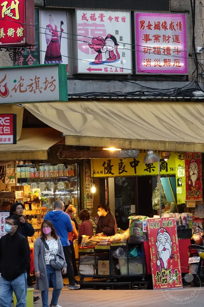 Ein belebter Ausschnitt der Dihua Street in Taipeh, Taiwan, zeigt traditionelle Geschäfte mit bunten Schildern und Auslagen von Waren. Mehrere Personen, einige mit Masken, navigieren durch die Gasse und verdeutlichen das geschäftige Treiben. Die Szene vermittelt eine lebendige und authentische Atmosphäre des städtischen Lebens.