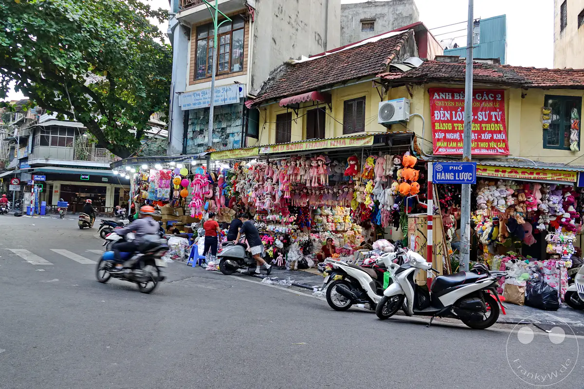 Straßenszene im Old Quarter von Hanoi, Vietnam, wo ein belebter Markt mit Spielzeug und Plüschtieren zum Verkauf steht. Motorroller passieren die bunte und lebhafte Gasse, die das tägliche Leben in der Stadt widerspiegelt. Die Atmosphäre ist geschäftig und typisch für die lebendigen Märkte dieser historischen Gegend.