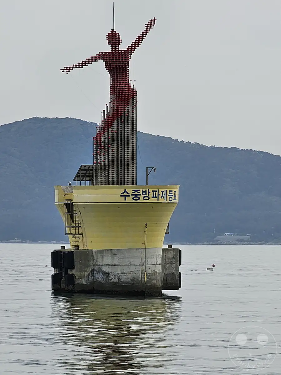 Die "Red Lady"-Skulptur erhebt sich auf einem schwimmenden Fundament im Wasser vor der Küste Südkoreas. Das imposante Bauwerk, das an die Wellenbrecherin von Haeundae Beach erinnert, thront über dem ruhigen Meer. Die maritime Umgebung und die beeindruckende Kunstinstallation schaffen eine atmosphärische und einzigartige Szenerie.
