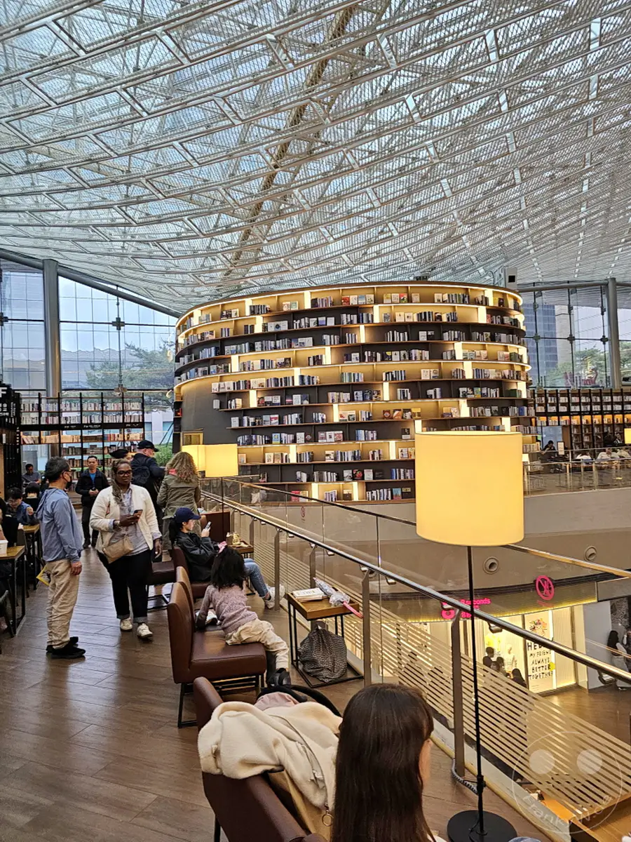 Die Starfield Library in Seoul, Südkorea, präsentiert sich mit einer beeindruckenden kreisförmigen Architektur, die von hohen Bücherregalen dominiert wird. Menschen sitzen und gehen durch die weitläufige Bibliothek, die unter einem futuristischen Glasdach liegt. Die Atmosphäre ist ruhig und einladend, ideal zum Lesen und Entspannen inmitten von Büchern.