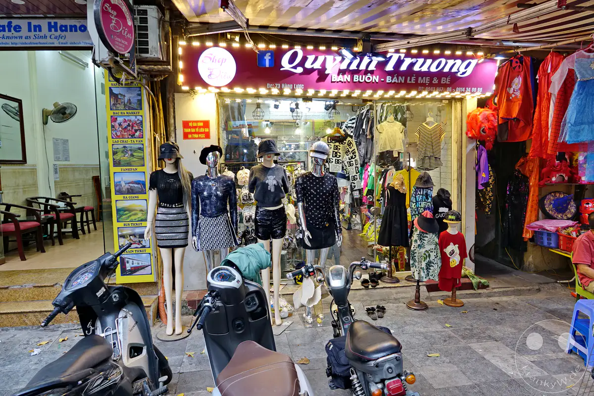 Die belebte Straße im Old Quarter von Hanoi, Vietnam, präsentiert ein lebhaftes Bekleidungsgeschäft mit Schaufensterpuppen und Kleiderständern, die eine Auswahl an Modeartikeln zur Schau stellen. Motorroller stehen auf dem Gehsteig, während Sitzgelegenheiten im Freien die Szene der geschäftigen Stadt ergänzen. Bunte Schilder und Hängeleuchten tragen zur lebendigen Atmosphäre dieses städtischen Umfelds bei.