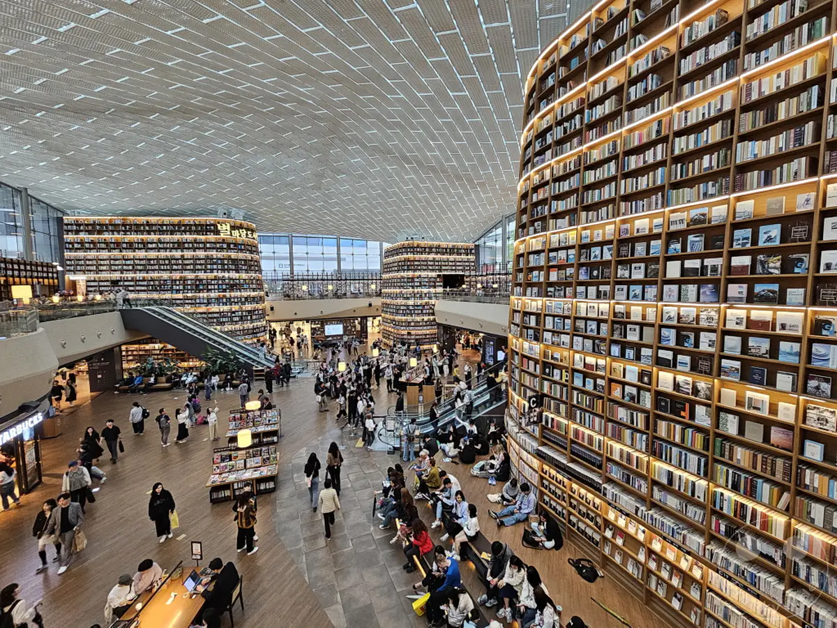 Die Starfield Library in Seoul, Südkorea, ist ein beeindruckendes Beispiel moderner Architektur und des Designs von Bibliotheken. Umgeben von riesigen, beleuchteten Bücherregalen, die vom Boden bis zur Decke reichen, bewegen sich zahlreiche Besucher durch den weitläufigen Raum. Die Atmosphäre ist lebhaft, während Menschen stöbern, sitzen und die einzigartige Umgebung genießen.