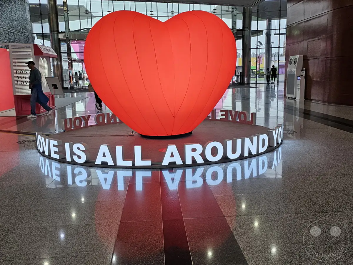 Eine riesige rote Herzskulptur steht zentral in der Starfield Coex Mall in Gangnam, Seoul, Südkorea. Die Installation ist von leuchtenden Buchstaben umgeben, die den Satz "LOVE IS ALL AROUND YOU" bilden und eine festliche Atmosphäre schaffen. Das glänzende Fußbodendesign spiegelt die Skulptur und die Beleuchtung wider und verstärkt die visuelle Wirkung.