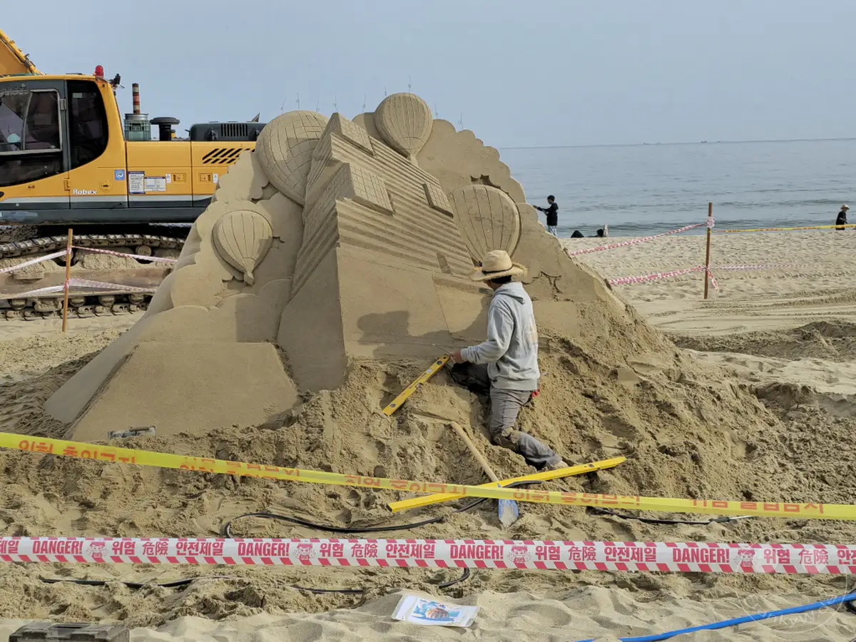 Ein Künstler in Südkorea, am Haeundae Beach in Busan, arbeitet an einer beeindruckenden Sandskulptur mit Heißluftballons und architektonischen Elementen. Ein Bagger steht im Hintergrund, während Absperrbänder die Baustelle sichern. Die Szene vermittelt einen Eindruck von kreativer Arbeit unter freiem Himmel am Meer.