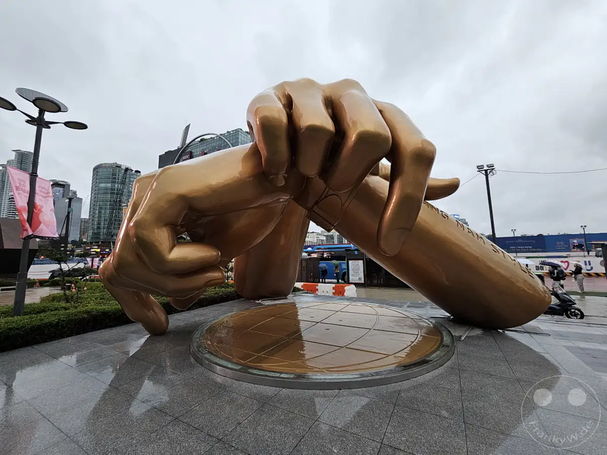 Eine goldene, übergroße Skulptur, die die Geste des Gangnam Style darstellt, steht im Freien unter einem bewölkten Himmel. Regen spiegelt sich auf dem nassen Boden und der metallischen Oberfläche des Kunstwerks, was eine melancholische, aber dennoch imposante Atmosphäre erzeugt. Die umliegende städtische Landschaft mit Wolkenkratzern bildet einen modernen Kontrast zu der ikonischen Skulptur in Seoul, Südkorea.