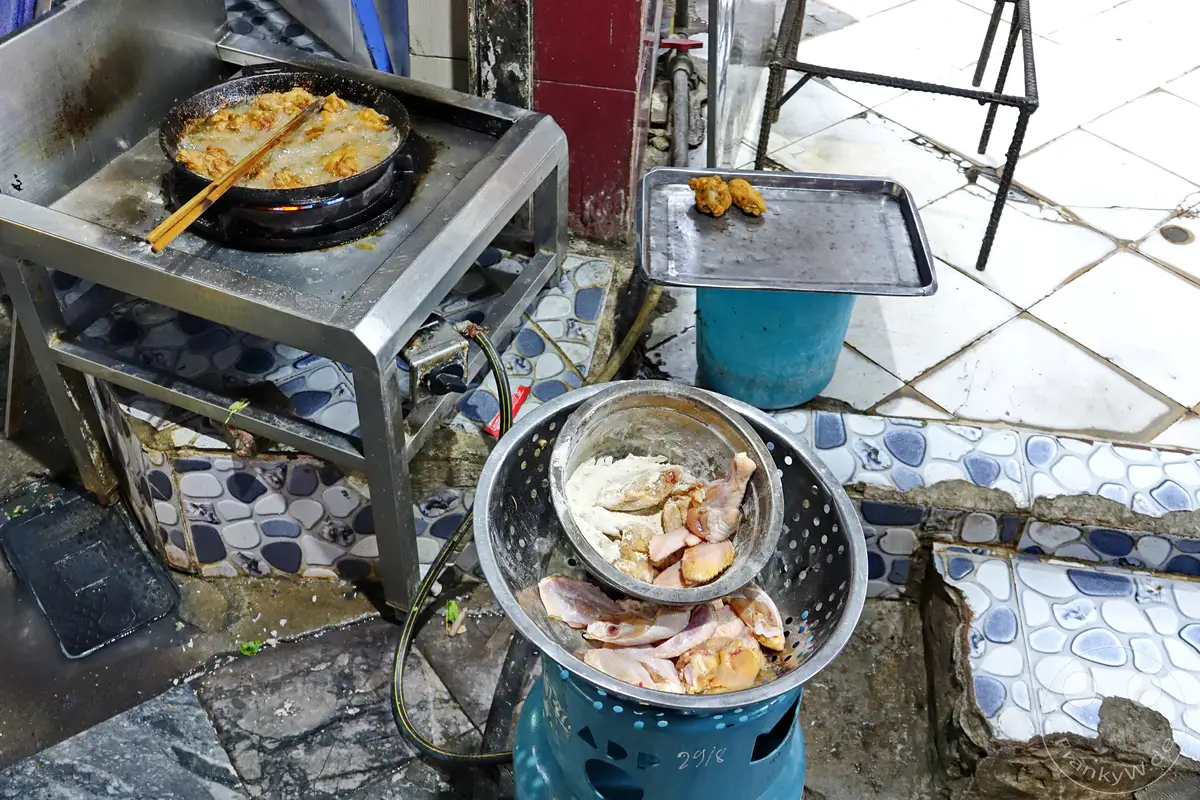 In einer Gasse des vietnamesischen Hanoi, in der Altstadt, werden frische Hähnchenflügel paniert und über offenem Feuer in heißem Öl frittiert. Die Szene vermittelt die authentische Atmosphäre des lokalen Streetfoods, wo die Zubereitung von Speisen oft unter freiem Himmel stattfindet.