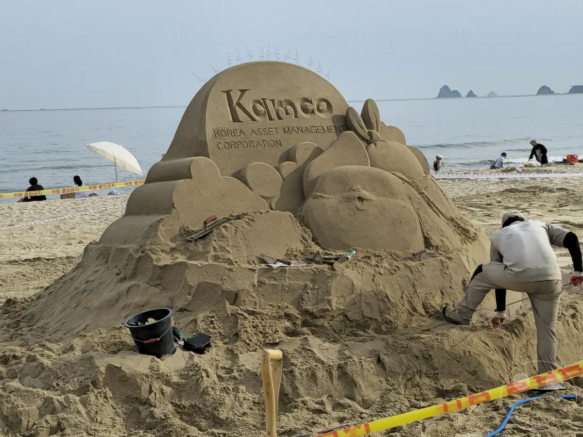 Am Haeundae Beach in Busan, Südkorea, wird eine beeindruckende Sandskulptur fertiggestellt, die das Logo von "Korea Asset Management Corporation" zeigt. Mehrere Personen arbeiten an der detailreichen Kreation, während andere im Hintergrund am Meer entspannen und die sommerliche Atmosphäre genießen. Die Szene vermittelt eine friedliche und kreative Stimmung an einem sonnigen Strandtag.
