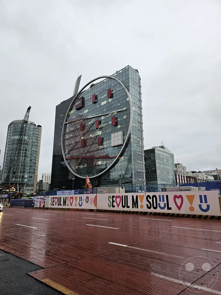 Ein imposantes modernes Gebäude mit einer kreisförmigen Fassade dominiert die Skyline von Seoul, Südkorea. An der Baustelle vor dem Gebäude weht die Botschaft "Seoul My Soul" auf einer bedruckten Lärmschutzwand. Der nasse, rote Straßenbelag im Vordergrund reflektiert das graue, wolkenverhangene Himmelslicht und unterstreicht die städtische Atmosphäre.