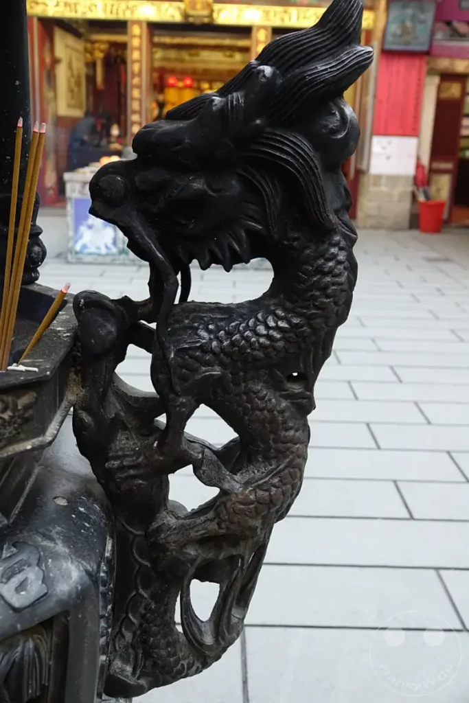 Ein detailliertes Drachenornament aus dunklem Metall ziert einen Räuchergefäß vor einem traditionellen Gebäude in Taipeh, Taiwan. Mehrere Räucherstäbchen sind angezündet und verströmen ihren Duft in der stimmungsvollen Atmosphäre des Dihua Street Tempelbezirks. Die Szene vermittelt einen Eindruck von lokaler Kultur und spiritueller Praxis.