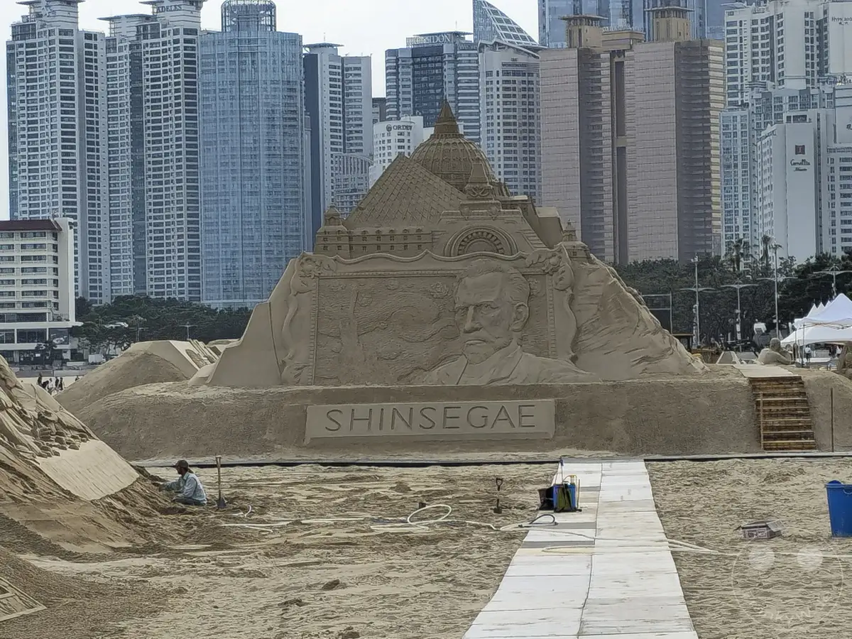 Eine beeindruckende Sandskulptur mit einem Porträt und architektonischen Elementen steht am Haeundae Beach in Busan, Südkorea. Hohe Gebäude bilden die Kulisse für diese beeindruckende Kunst am Strand. Die Szene vermittelt eine Atmosphäre der Kreativität und der sommerlichen Freude.
