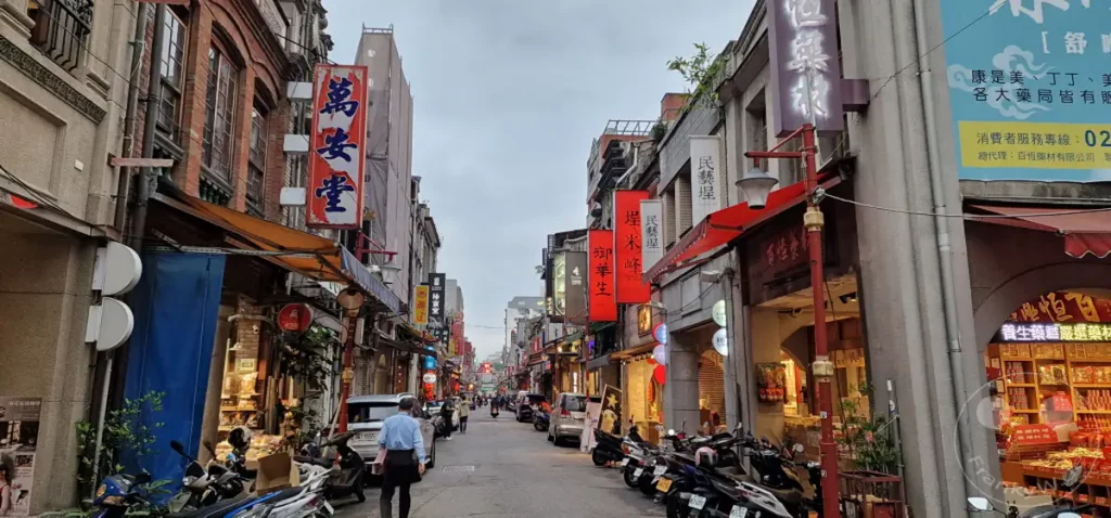 Die belebte Dihua Street in Taipeh, Taiwan, präsentiert sich mit traditionellen Geschäften und bunten Schildern unter einem bewölkten Himmel. Motorroller säumen die Gasse, während Menschen ihren täglichen Besorgungen nachgehen und die Atmosphäre dieses historischen Viertels durchdringt. Die Architektur und die Geschäfte spiegeln eine Mischung aus Vergangenheit und Gegenwart in dieser geschäftigen asiatischen Metropole wider.