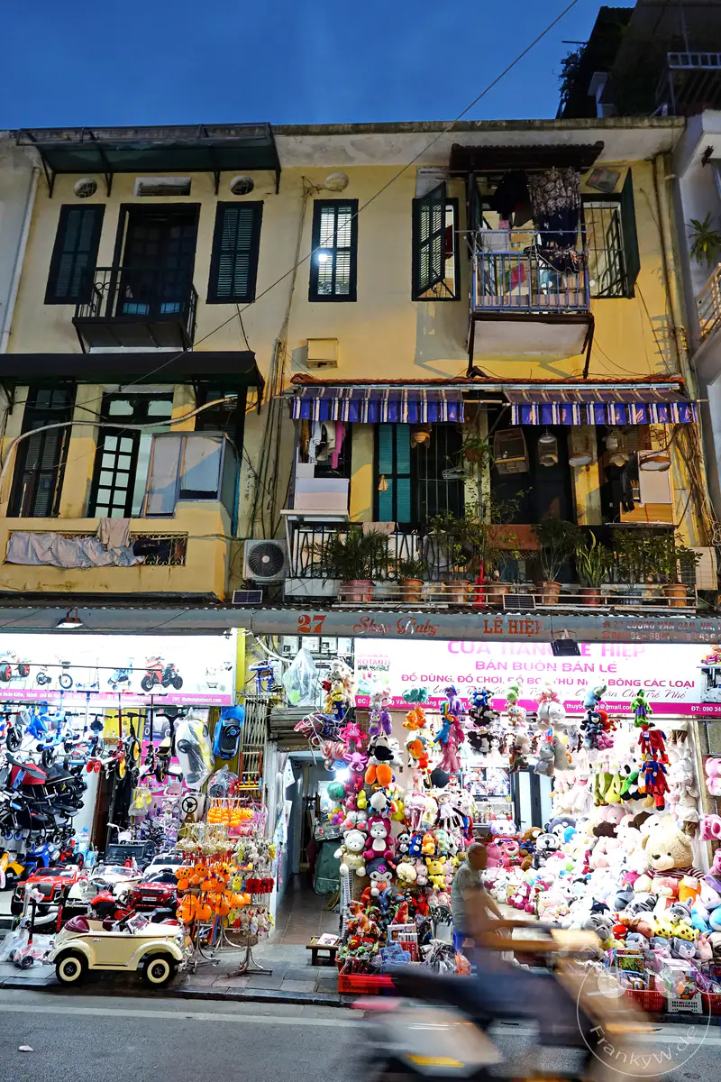 Die Lichter eines Spielzeugladens in Hanois Altstadt in Vietnam erhellen die Nacht und präsentieren eine bunte Auswahl an Stofftieren und Spielzeugautos. Die Fassade des Gebäudes mit mehreren Etagen und Balkonen gibt Einblick in das alltägliche Leben. Die belebte Straße mit vorbeifahrenden Motorrollern schafft eine lebendige und geschäftige Atmosphäre.