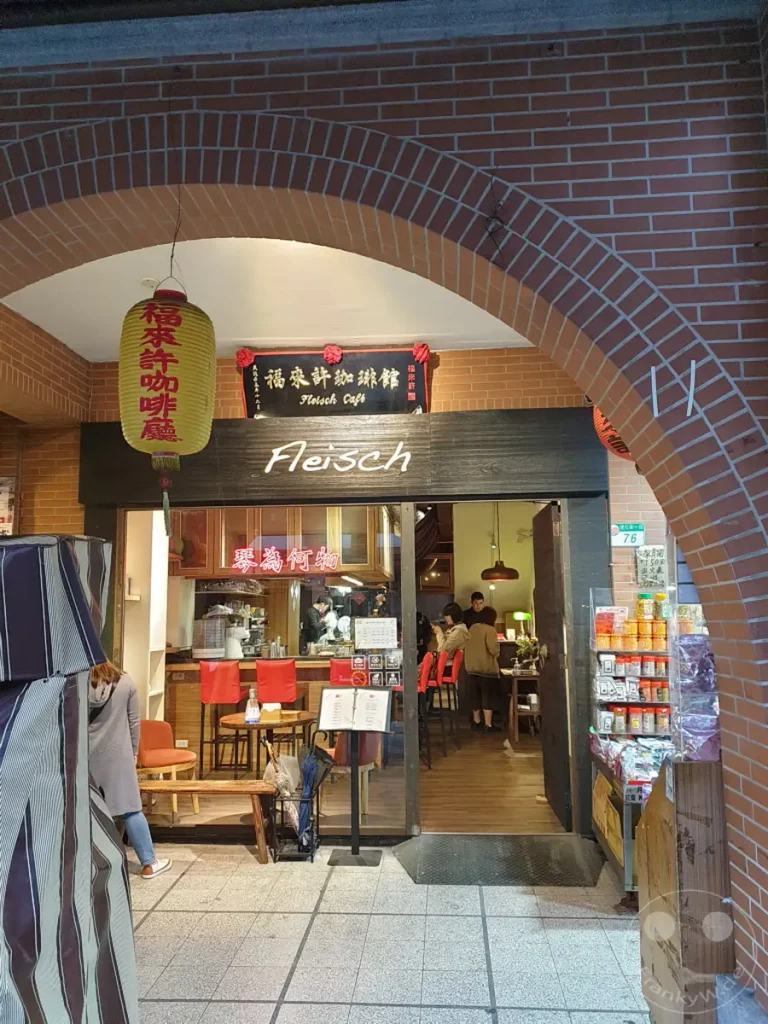 Der Eingangsbereich eines Cafés in Taipeh, Taiwan, wird von einer Ziegelsteinfassade eingerahmt. Eine gelbe Lampionschale hängt über dem Eingang zum „Fleisch Cafe“, das durch seine Holzverkleidung und das beleuchtete Innere einladend wirkt. Kleine Tische und rote Barhocker laden zum Verweilen ein, während im Hintergrund geschäftiges Treiben zu erkennen ist.