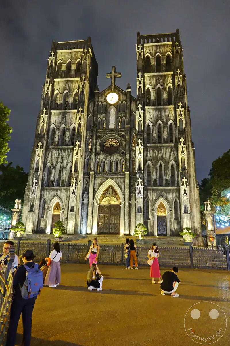 Die St. Joseph's Cathedral in Hanoi, Vietnam, erstrahlt bei Nacht in festlicher Beleuchtung. Menschenmengen versammeln sich vor dem imposanten neugotischen Bauwerk, um seine Architektur zu bestaunen und die Atmosphäre einzufangen. Die beleuchtete Fassade und die Silhouette der Kirche gegen den dunklen Nachthimmel schaffen eine mystische und eindrucksvolle Szenerie.