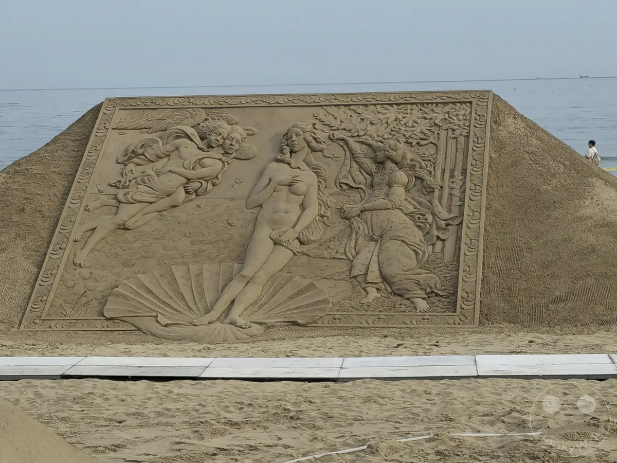 Eine beeindruckende Sandskulptur am Haeundae Beach in Busan, Südkorea, stellt eine Szene dar, die an die Geburt der Venus erinnert. Die detailreiche Darstellung im Freien vor der Kulisse des Meeres erzeugt eine ruhige und künstlerische Atmosphäre. Kleinere Figuren und ein Muschelfundament ergänzen das Hauptmotiv der Sandkunst.