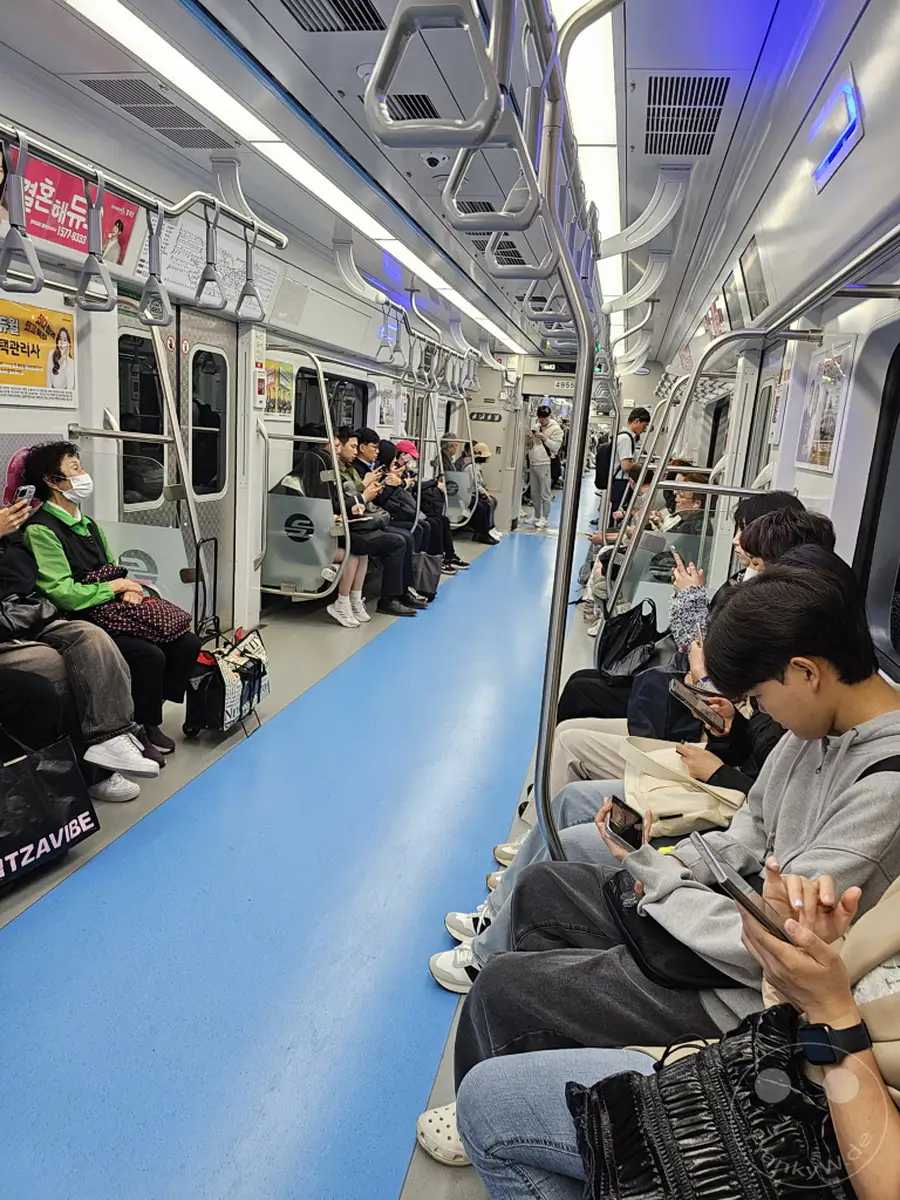 Eine überfüllte U-Bahn in Seoul, Südkorea, mit vielen Passagieren, die auf den blauen Sitzen und dem Gang sitzen. Die meisten Menschen schauen auf ihre Handys und wirken in ihre eigenen Welten versunken. Die Szene vermittelt ein Gefühl des geschäftigen Alltags in der südkoreanischen Hauptstadt.