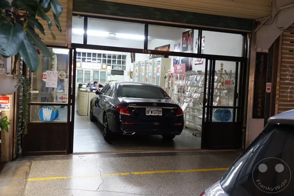 Ein dunkles Mercedes-Benz E200 steht in einer offenen Garage, die zu einem Geschäft führt. Regale mit Produkten sind im Hintergrund sichtbar, und ein Schild weist auf die Nummer 86 hin. Die Szene vermittelt einen Eindruck des geschäftigen Treibens auf Taipehs Dihua Street.