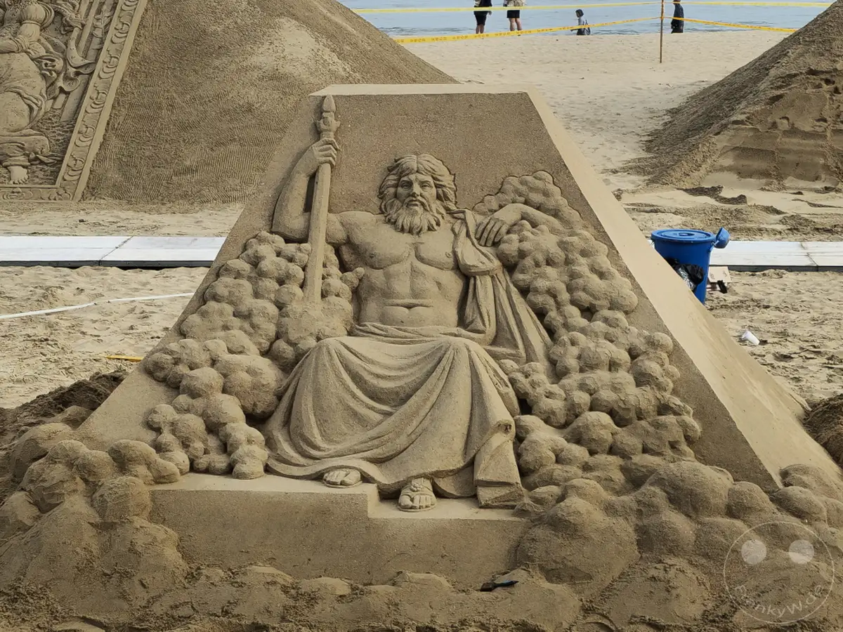 Ein imposantes Sandskulpturen-Kunstwerk am Haeundae Beach in Busan, Südkorea, stellt eine sitzende Götterfigur mit Bart und Zepter dar. Umgeben von Wolken oder stürmischer See spiegelt die Skulptur die Kraft und Erhabenheit der dargestellten mythologischen Figur wider. Die Darstellung in Sandform erzeugt eine beeindruckende und vergängliche Kunstform am Strand.