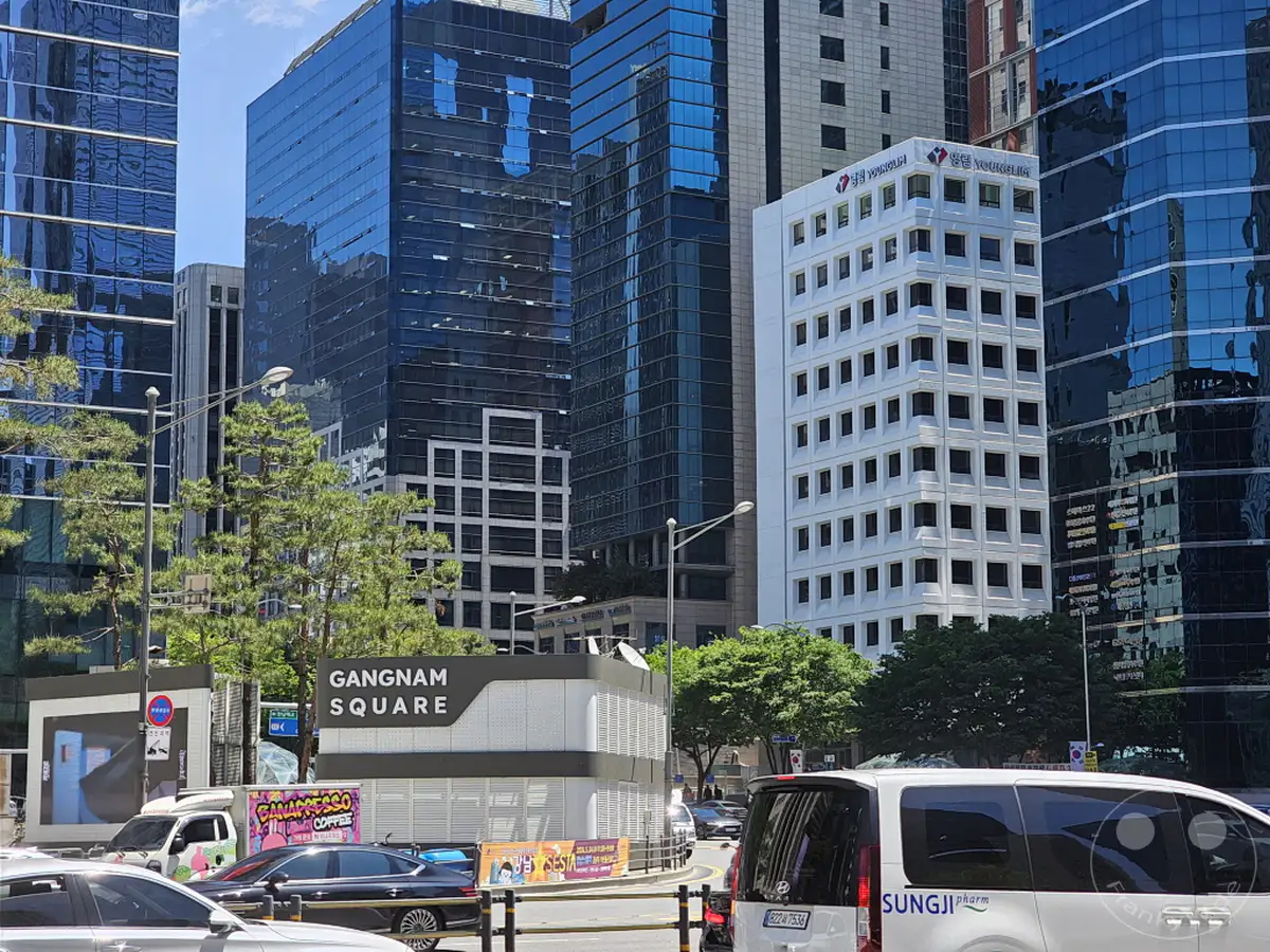 Moderne Glasgebäude dominieren das Stadtbild von Seoul, Südkorea, im geschäftigen Viertel Gangnam Square. Ein weißes Gebäude mit vielen Fenstern steht im Kontrast zu den umliegenden dunklen Glasbauten. Bäume und Fahrzeuge auf der Straße verleihen der städtischen Kulisse Lebendigkeit.