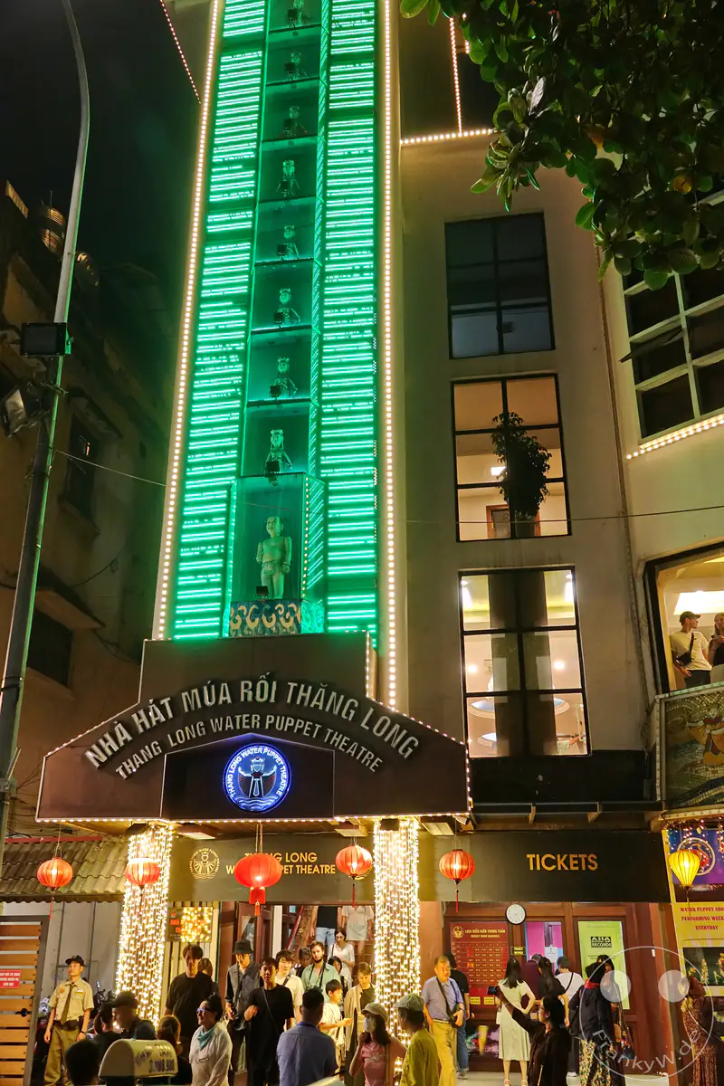 Das Thang Long Water Puppet Theatre in Hanoi, Vietnam, erstrahlt nachts in bunten Lichtern und lädt zu einer Vorstellung ein. Eine Gruppe von Menschen wartet auf Einlass und imposante traditionelle Figuren zieren die Fassade des Gebäudes. Die beleuchtete Architektur und die bunten Dekorationen schaffen eine lebendige und einladende Atmosphäre.