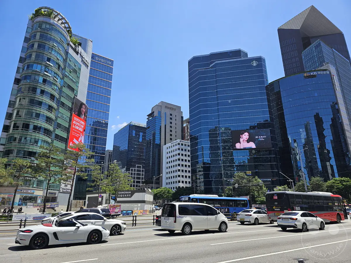 Ein Blick auf die beeindruckende Skyline von Gangnam Square in Seoul, Südkorea, an einem sonnigen Tag. Moderne Wolkenkratzer prägen das Stadtbild, während auf der mehrspurigen Straße reger Fahrzeugverkehr herrscht. Die Szene vermittelt ein Gefühl von urbaner Dynamik und Fortschritt.
