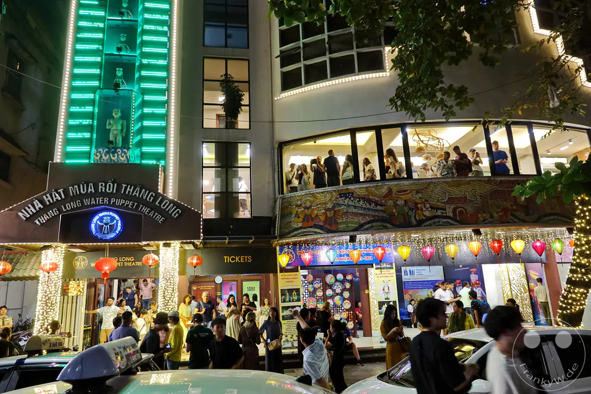 Eine lebhafte Straßenszene in Hanoi, Vietnam, am Abend vor dem Thang Long Water Puppet Theatre. Bunte Lichterketten und Laternen beleuchten die Fassade, während Menschen sich vor dem Eingang versammeln und die bunte Atmosphäre genießen. Das Bild fängt die kulturelle Energie und das geschäftige Treiben der vietnamesischen Hauptstadt ein.