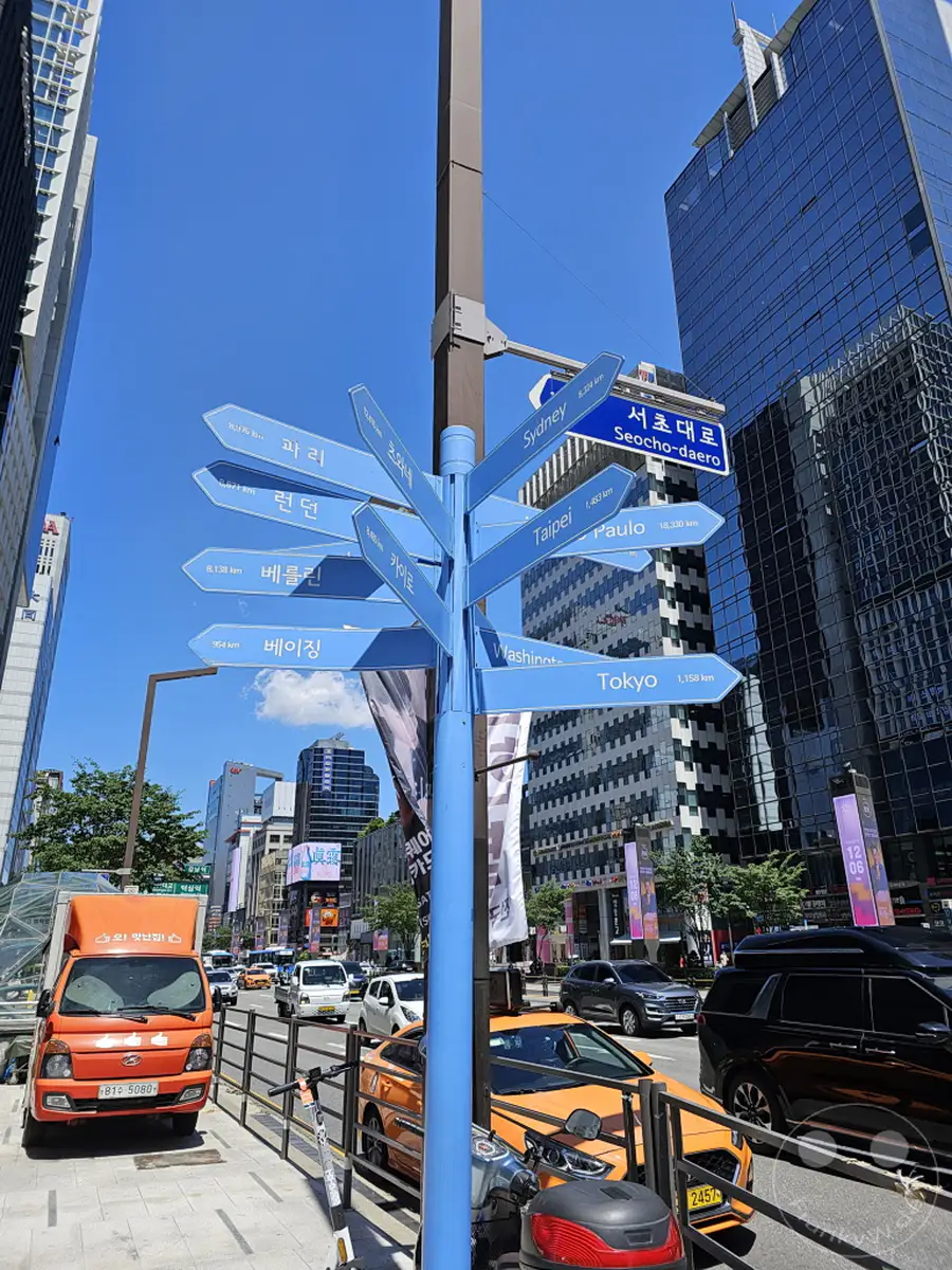 Ein blauer Wegweiser in Seoul, Südkorea, zeigt die Entfernungen zu verschiedenen internationalen Städten wie Sydney, Tokio, Washington und Berlin. Umgeben von modernen Wolkenkratzern und belebten Straßen, spiegelt die Szene die globale Vernetzung der Metropole wider. Die klare blaue Himmelkulisse unterstreicht die städtische Lebendigkeit und die geografische Ausrichtung der Stadt.