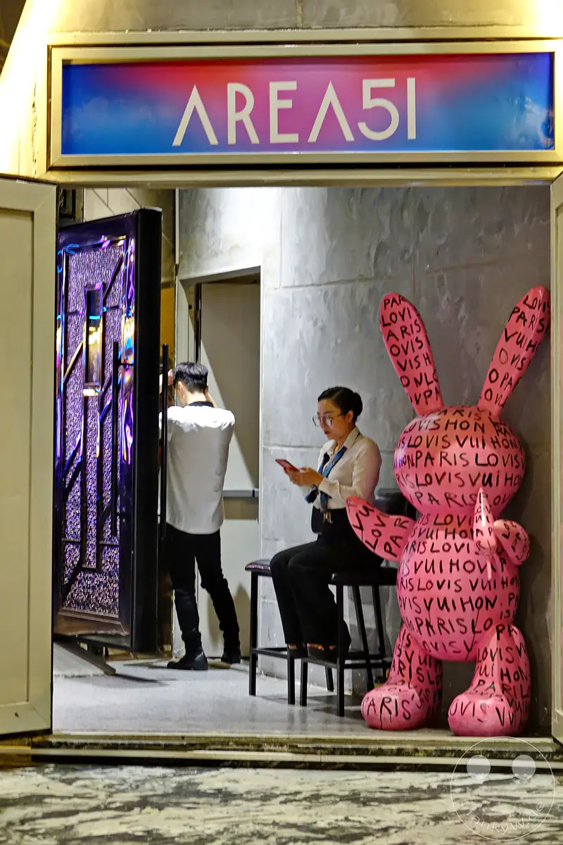 Eine Szene in Hanoi, Vietnam, mit der leuchtenden AREA51-Beschilderung über einem Eingangsbereich, der von einem Mann und einer Frau belebt wird. Auffällig ist eine große rosa Hasenfigur, die mit Louis Vuitton-Schriftzügen dekoriert ist und die urbane Atmosphäre der Nacht ergänzt.