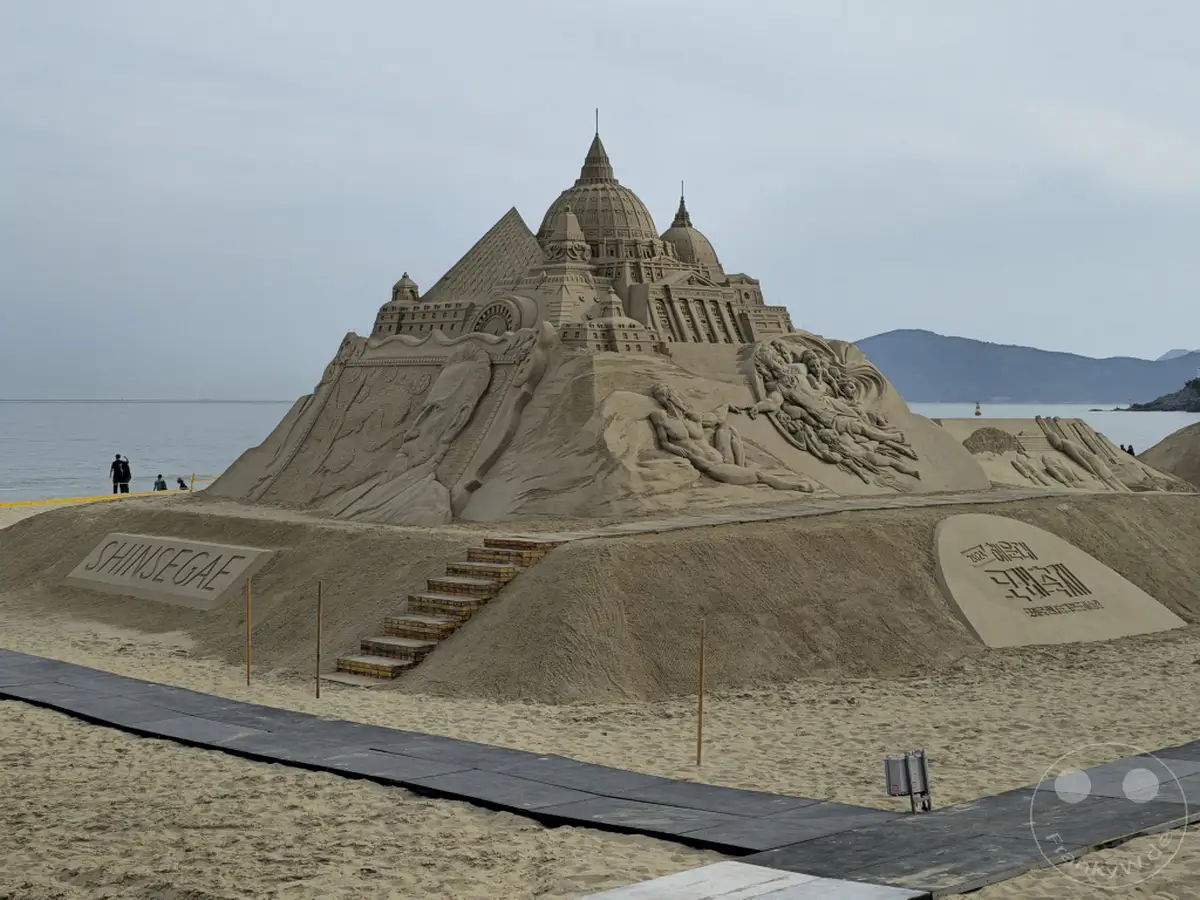 Eine beeindruckende Sandskulptur, die an eine antike Stadt erinnert, steht am Haeundae Beach in Busan, Südkorea. Die filigranen Details und die Größe des Kunstwerks ziehen die Blicke auf sich und spiegeln die Kreativität der Künstler wider. Die leicht bewölkte Himmel und das ruhige Meer schaffen eine friedliche Atmosphäre für diese temporäre Strandkunst.