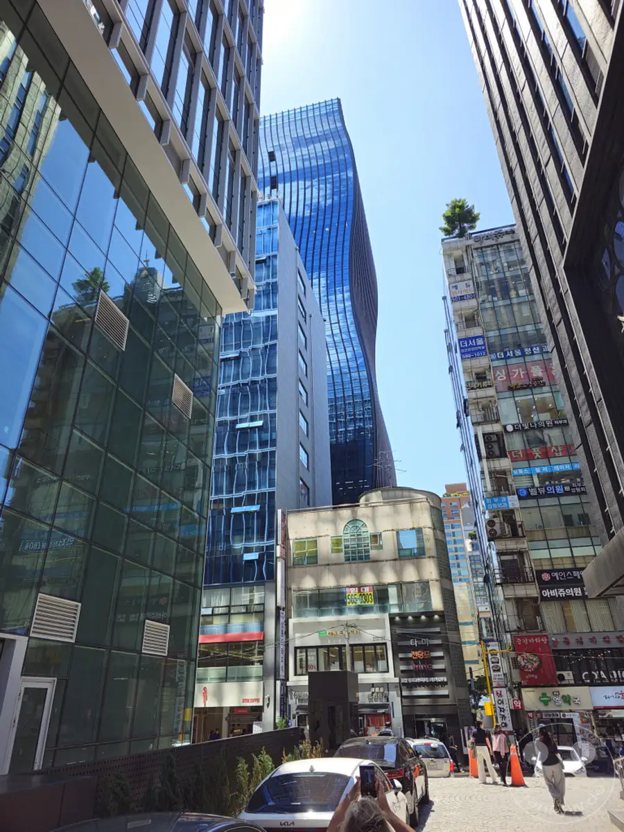 In Seoul, Südkorea, erhebt sich eine beeindruckende Skyline aus modernen Wolkenkratzern, die den pulsierenden Gangnam Square dominieren. Glänzende Glasfassaden spiegeln den strahlend blauen Himmel wider und erzeugen ein dynamisches, futuristisches Stadtbild. Das geschäftige Treiben der Metropole ist sowohl in der Architektur als auch in den belebten Straßen spürbar.