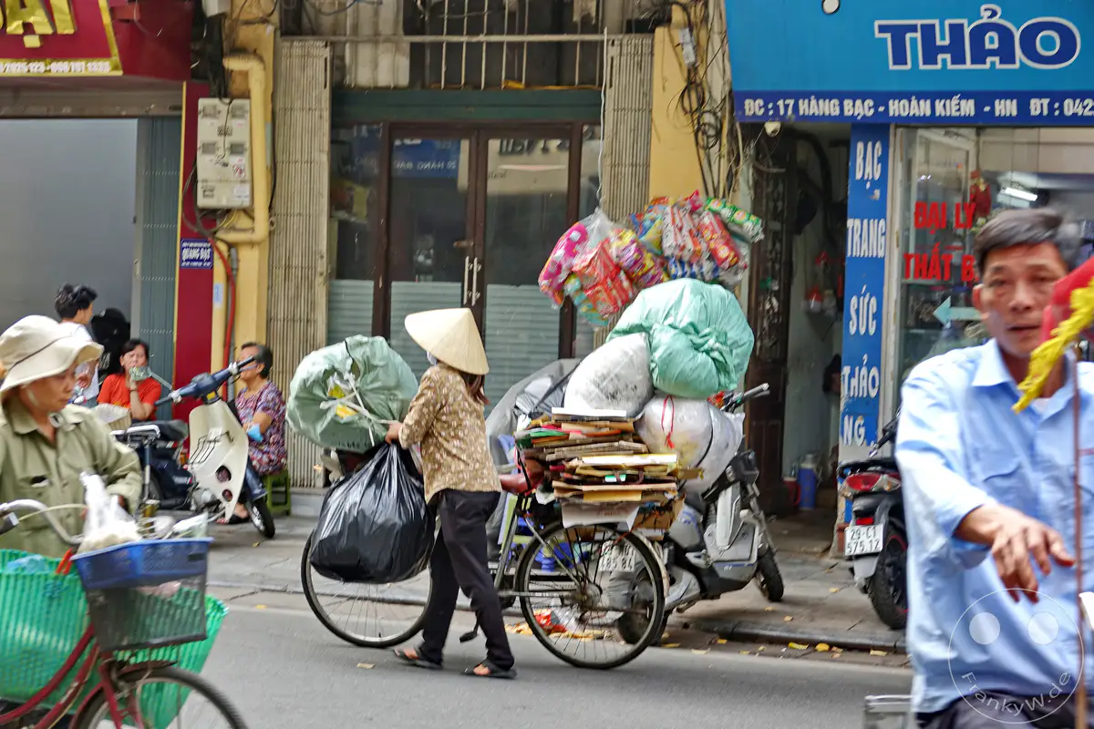 In einer geschäftigen Gasse des Old Quarter in Hanoi, Vietnam, transportiert eine Frau mit einem traditionellen Kegelhut und einem Fahrrad mehrere große Säcke und Pakete. Passanten und Motorroller beleben die Szene und vermitteln einen lebendigen Eindruck des täglichen Treibens im Herzen der Stadt. Die Atmosphäre ist geprägt von Geschäftigkeit und dem authentischen Charakter einer asiatischen Metropole.