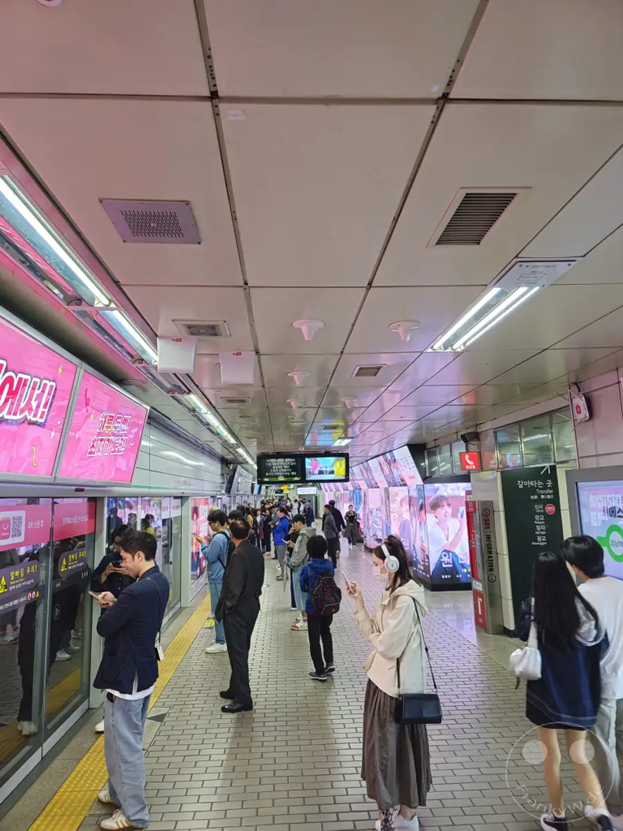 Menschen warten auf dem Bahnsteig einer belebten U-Bahn-Station in Seoul, Südkorea. Leuchtende Werbetafeln säumen die Plattform und spiegeln sich in den geschlossenen Türen des einfahrenden Zuges. Die Szene vermittelt das geschäftige Treiben des täglichen Lebens in der südkoreanischen Hauptstadt.