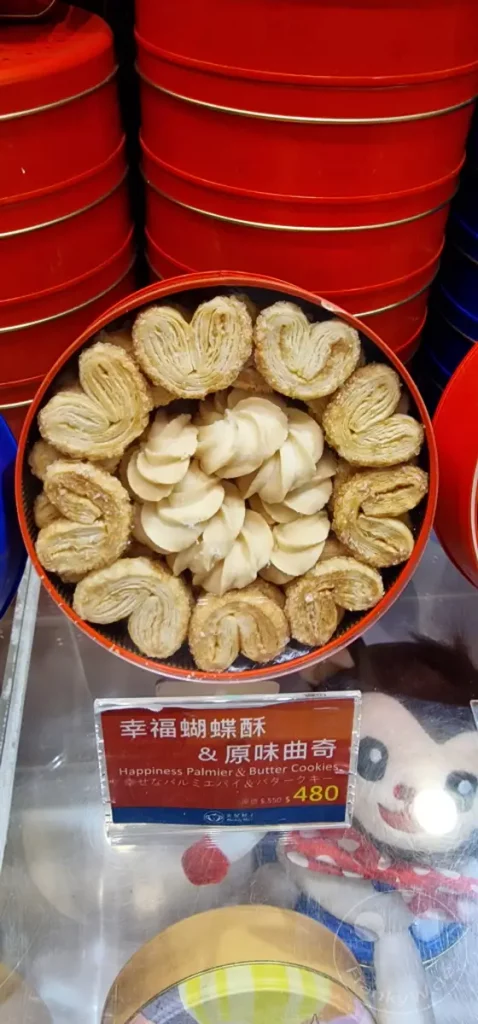 In Taipeh, Taiwan, präsentiert sich eine rote Blechdose voller verschiedener Kekse, darunter die zarten Palmiers und cremige Butterkekse. Das Etikett informiert auf Chinesisch und Englisch über 'Happiness Palmier & Butter Cookies' und einen Preis von 480. Im Hintergrund sind weitere rote Dosen und eine Stoffpuppe zu sehen, was auf ein geschäftiges Markt- oder Ladenszenario in der Dihua Street hindeutet.