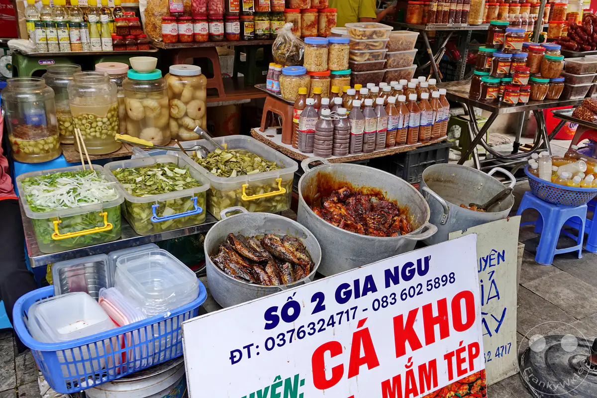 Auf dem lebhaften Markt im Old Quarter – die Altstadt von Hanoi, Vietnam, werden verschiedene fermentierte Fischsaucen und eingelegtes Gemüse zum Verkauf angeboten. Mehrere große Behälter enthalten zubereitete Fischgerichte und frische Zutaten, die die kulinarische Vielfalt des Landes widerspiegeln. Die Szene vermittelt eine authentische Atmosphäre eines lokalen Markttages mit einer Fülle von Lebensmittelangeboten.