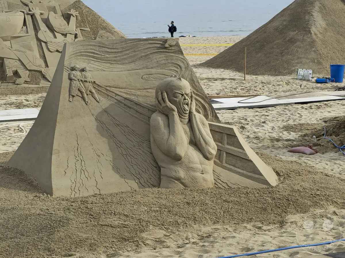 Eine monumentale Sandskulptur am Haeundae Beach in Busan, Südkorea, stellt eine dramatische Szene dar, die menschliches Leid und Verzweiflung symbolisiert. Die detailreiche Darstellung von Figuren, die mit ihren Händen die Ohren bedecken und mit weit geöffnetem Mund schreien, erzeugt eine kraftvolle und bewegende Atmosphäre. Im Hintergrund sind weitere Sandformationen und die Weite des Meeres zu sehen, was die Größe und den Umfang des Sandskulpturenfestivals unterstreicht.