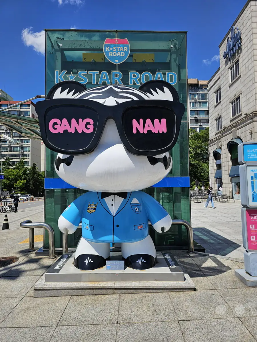 In Südkoreas Hauptstadt Seoul, auf der K-Star Road, steht eine auffällige Bärenfigur mit großen Sonnenbrillen, auf denen 'Gangnam' steht. Die bunte Skulptur ist Teil einer Attraktion, die die K-Pop-Kultur feiert. Die sonnige Atmosphäre und die städtische Kulisse laden zum Entdecken und Fotografieren ein.