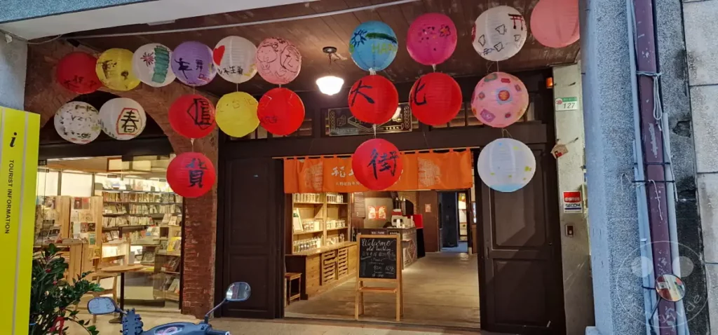Bunte Papierlaternen hängen über einer traditionellen Straße in Taipeh, Taiwan. Dahinter öffnet sich der Eingang zu einer Buchhandlung, die eine einladende und nostalgische Atmosphäre ausstrahlt. Die Szene kombiniert kulturelle Elemente mit der Ruhe eines Buchladens im Herzen der Dihua Street.