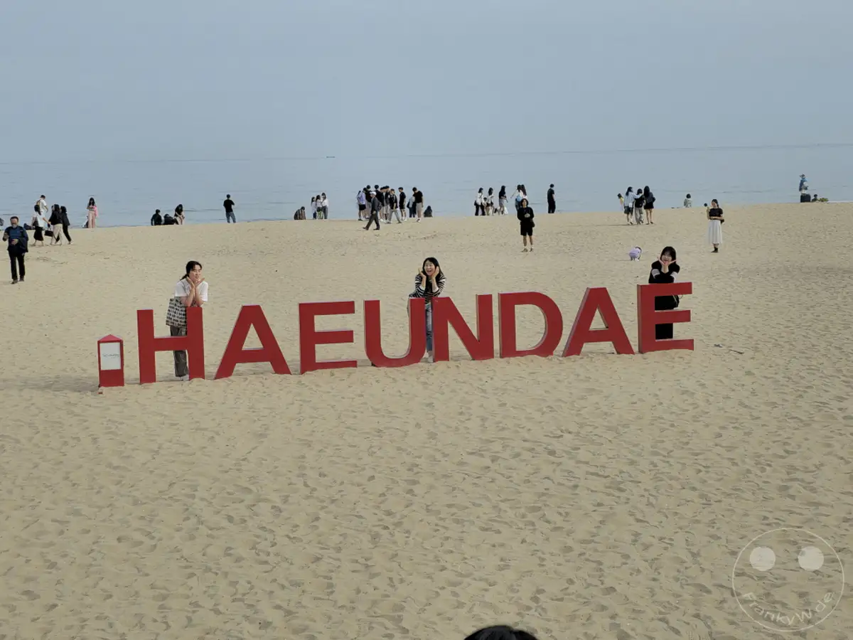 Am berühmten Haeundae Beach in Busan, Südkorea, stehen große rote Buchstaben mit der Aufschrift "HAEUNDAE" im Sand. Im Hintergrund tummeln sich zahlreiche Menschen am Wasser und genießen die Sonne. Die friedliche Atmosphäre am Strand lädt zum Entspannen und Erholen ein.