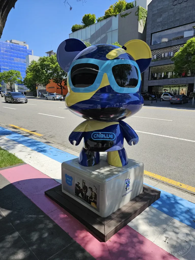 Eine bunte Skulptur eines stilisierten Bären steht auf der Star Road in Gangnam, Seoul, Südkorea. Die Skulptur ist mit einem auffälligen Design in Blau- und Gelbtönen versehen und trägt das Logo "CNBLUE". Die lebendige Gestaltung und die Umgebung deuten auf eine fröhliche und unterhaltsame Atmosphäre hin, die typisch für die K-Pop-Kultur ist.