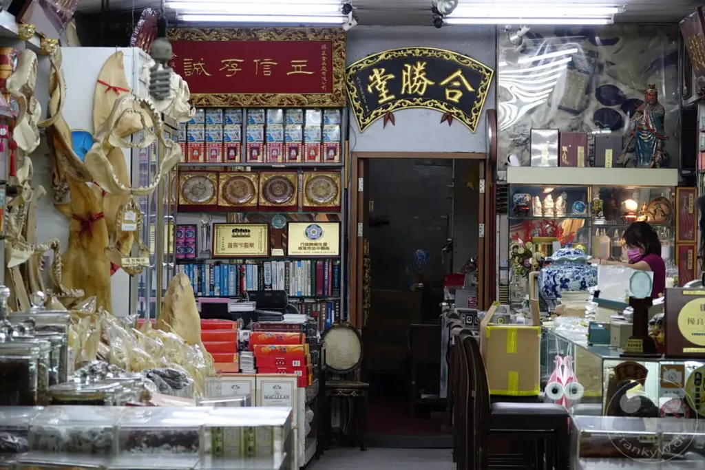 In einem traditionellen Laden in Taipehs Dihua Street werden verschiedene Heilkräuter, Trockenfischprodukte und andere traditionelle Waren angeboten. Die Regale sind dicht gefüllt und vermitteln eine Atmosphäre von reicher Geschichte und kulturellem Erbe. Eine Verkäuferin im Hintergrund bedient eine Kundin, was den geschäftigen Alltag des Ladens unterstreicht.