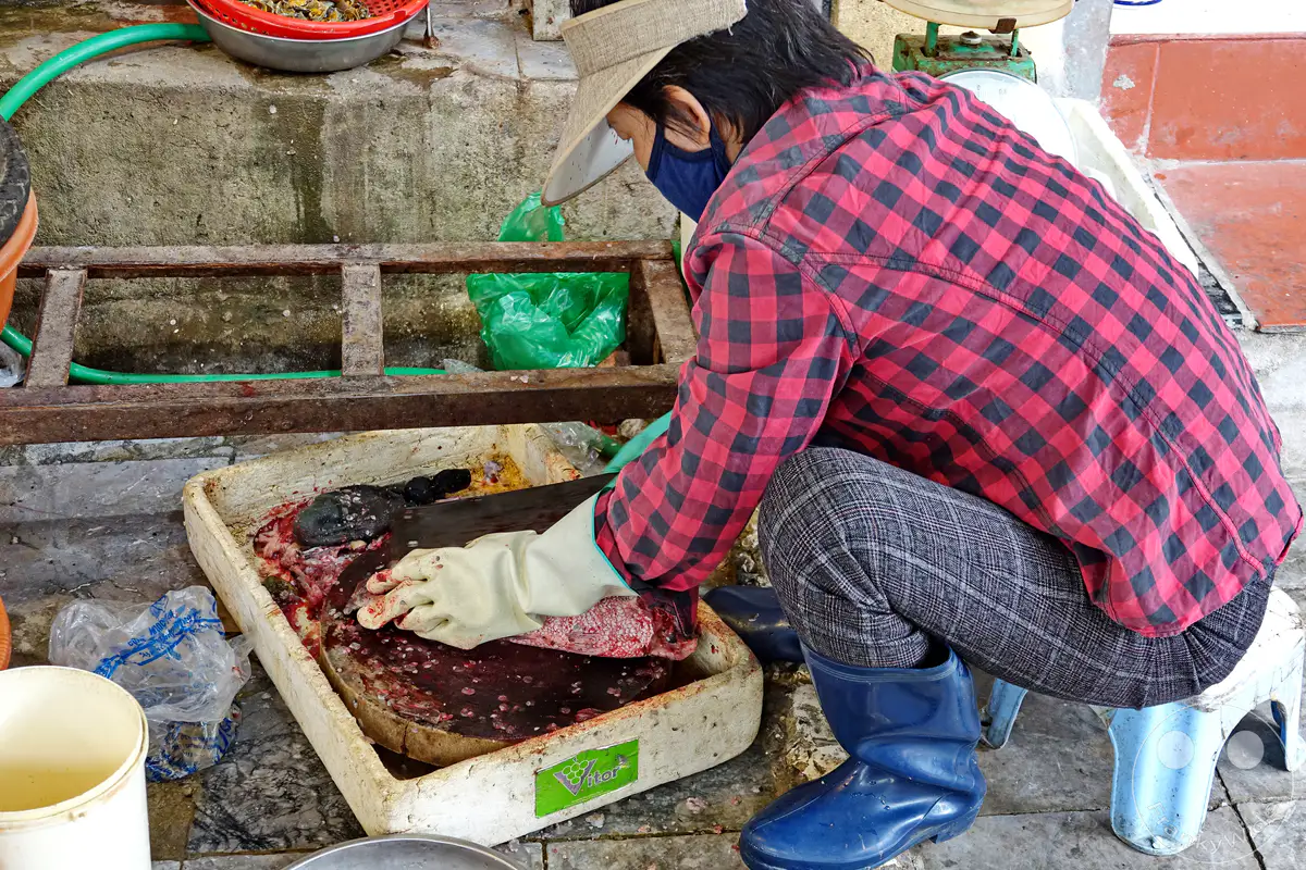 In Hanoi, Vietnam, verarbeitet ein Fischhändler im Old Quarter – die Altstadt – frischen Fisch auf einem Holzbrett. Das geschäftige Marktumfeld und die detaillierte Zubereitung vermitteln einen authentischen Einblick in das tägliche Leben und die kulinarische Kultur der Region. Die Szene ist geprägt von der praktischen Arbeitsweise unter freien Himmelbedingungen.