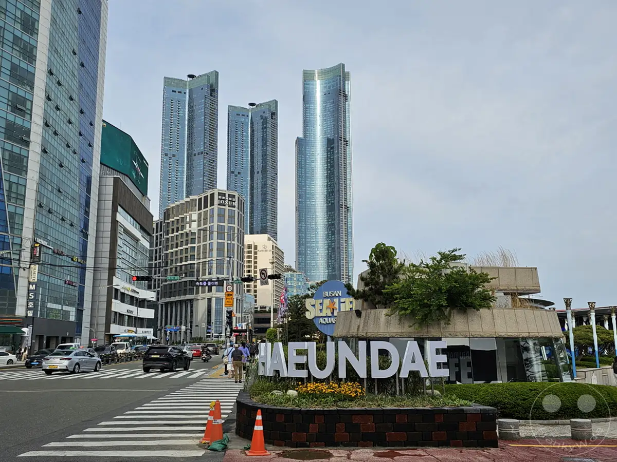 Die ikonische Skyline von Busan in Südkorea prägt das Bild mit modernen Wolkenkratzern und der bekannten Haeundae Beach Beschriftung. Autos passieren eine belebte Kreuzung, während Fußgänger die urbane Atmosphäre genießen. Das Gesamtbild vermittelt einen Eindruck von einer dynamischen und aufstrebenden Metropole.