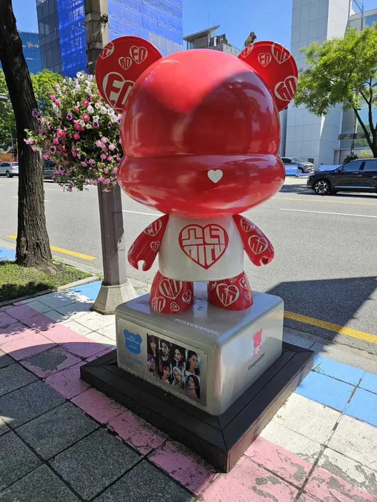 Eine knallrote Teddybär-Skulptur mit Herzen steht prominent auf einer "Star Road" in Seoul, Südkorea. Auf dem Sockel der Skulptur ist eine Gruppe von Bildern der K-Pop-Gruppe "Girls' Generation" abgebildet. Die Szene strahlt eine fröhliche und bunte Atmosphäre aus, die Fans und Touristen gleichermaßen anspricht.