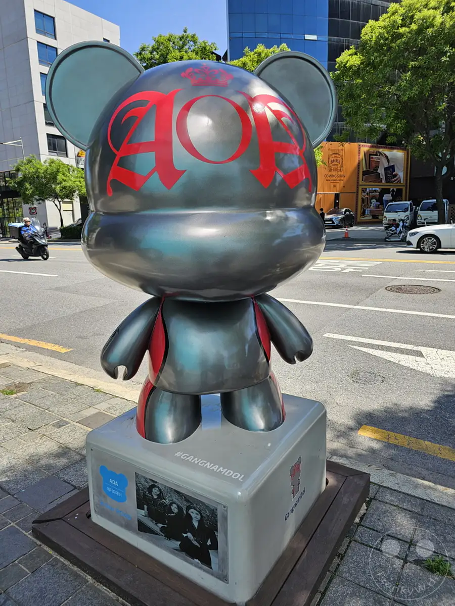 Eine humorvolle Bärenstatue mit der Aufschrift "AOA" und "GangnamDool" steht auf einem Sockel in Seoul, Südkorea. Ein Motorradfahrer fährt im Hintergrund vorbei, während in der Ferne ein Geschäft mit der Aufschrift "Coming Soon" zu sehen ist. Die strahlende Sonne und die gepflegten Bäume erzeugen eine lebhafte und urbane Atmosphäre.