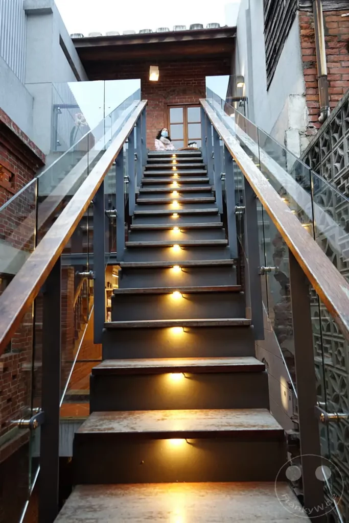 Eine beleuchtete Treppe führt in einem Gebäude in Taipeh, Taiwan, nach oben, flankiert von modernen Glas- und Metallgeländern sowie der Backsteinarchitektur der Dihua Street. Die dezente Beleuchtung der Stufen schafft eine warme und einladende Atmosphäre, während im Hintergrund eine Person auf den oberen Stufen sitzt. Das Zusammenspiel von Alt und Neu prägt das architektonische Bild dieser Szenerie.