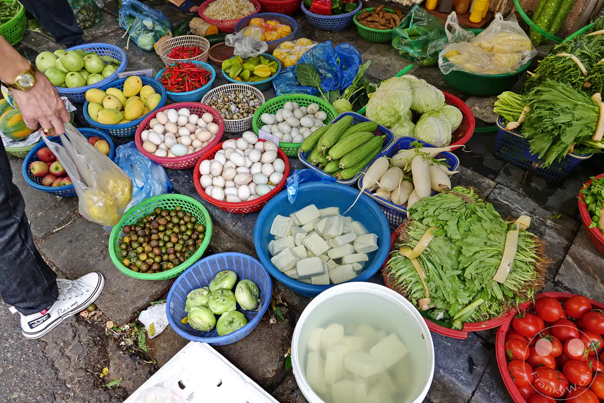 Bunte Vielfalt auf einem Markt in Hanois Altstadt in Vietnam. Verschiedene Körbe sind mit frischem Obst und Gemüse wie Tomaten, Rettichen, Wasserkastanien, Eiern und grünem Gemüse gefüllt. Die lebendige Atmosphäre spiegelt das tägliche Leben und die kulinarische Vielfalt des Landes wider.