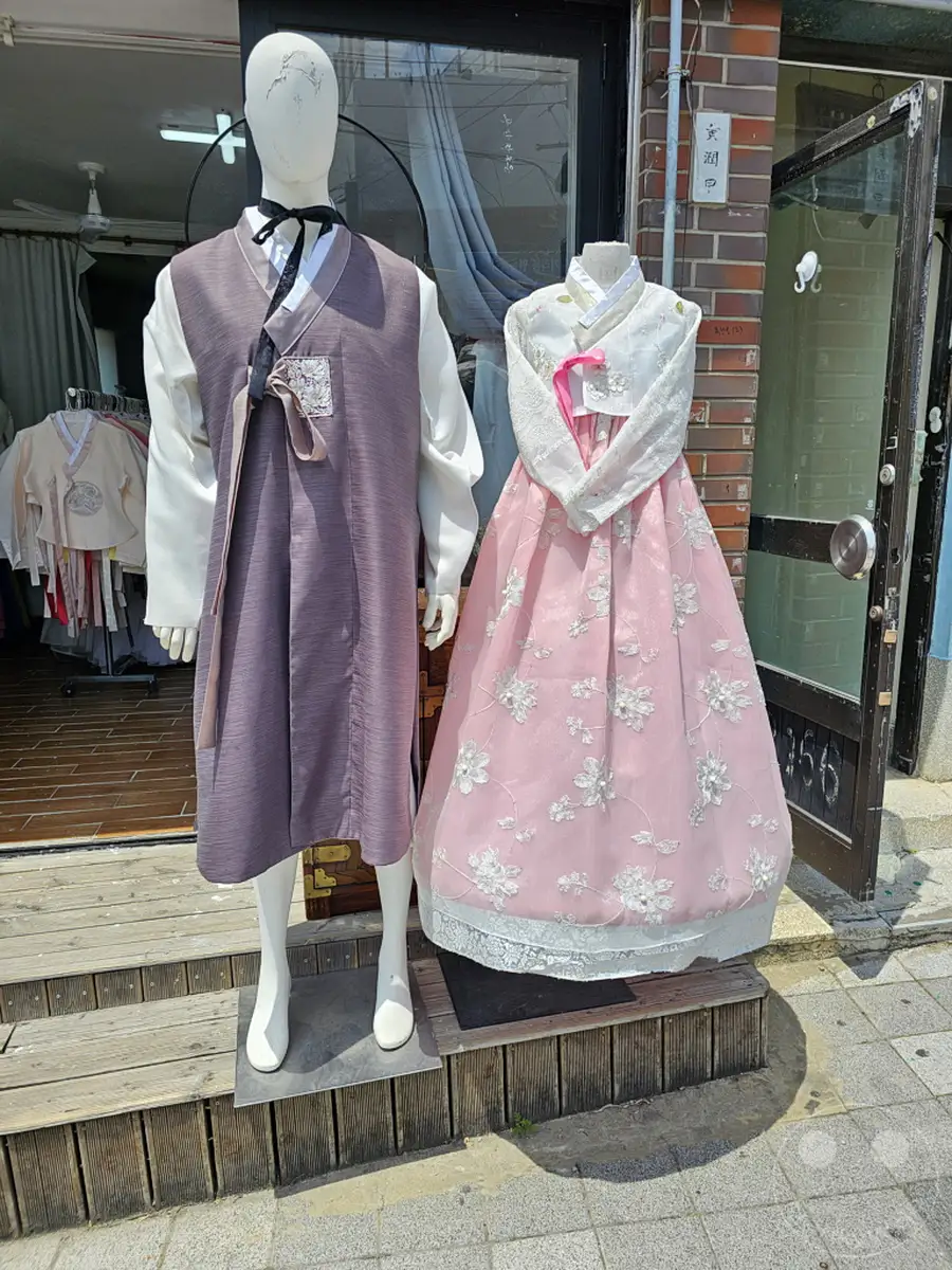 Auf den Stufen vor einem Geschäft im Culture Village Gamcheon in Südkorea werden zwei Mannequins in traditionellen Hanboks präsentiert. Ein männlicher Hanbok in Grau und Weiß steht neben einem femininen Modell in Rosa mit weißen Stickereien. Die Auslage schafft eine atmosphärische Verbindung zwischen zeitgenössischer Mode und der reichen kulturellen Geschichte des Ortes.